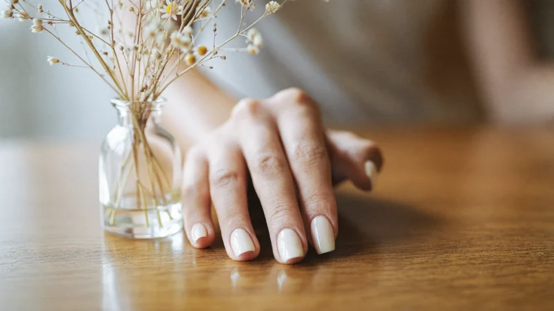 Illustration: Hand mit fertiger dezenter Maniküre neben Trockenblumen auf Holztisch